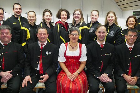 Gruppenfoto der Vorstandschaft. Vorne sitzen sechs Personen in traditioneller Tracht, hinten stehen neun Mitglieder in schwarzen Vereinsjacken mit gelben Streifen. Alle lächeln und blicken in die Kamera.