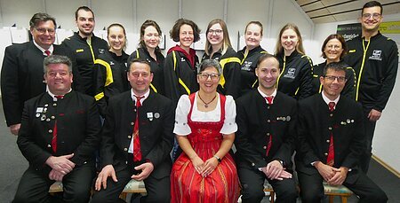 Gruppenfoto der Vorstandschaft. Vorne sitzen sechs Personen in traditioneller Tracht, hinten stehen neun Mitglieder in schwarzen Vereinsjacken mit gelben Streifen. Alle lächeln und blicken in die Kamera.